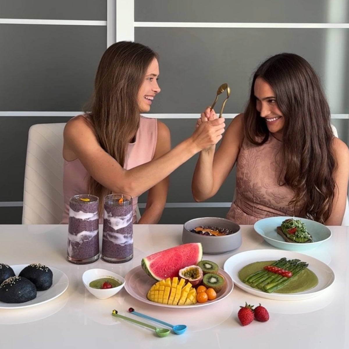 Two women drinking white and purple juice at a table surrounded by fruits and vegetables